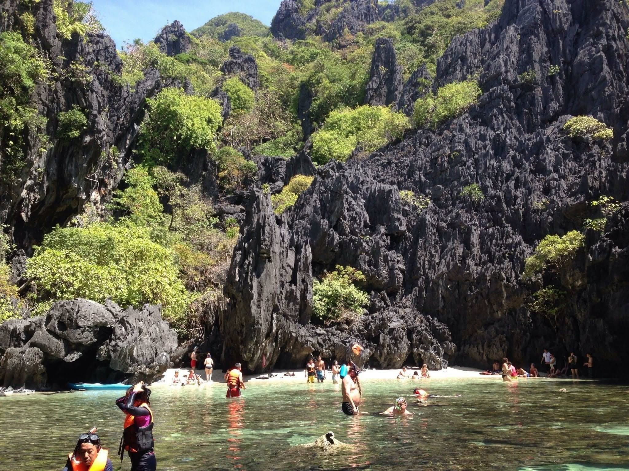 Swimmers and snorkellers in clear shallow water at a small beach enclosed by black limestone cliffs, on the Tour C route in El Nido
