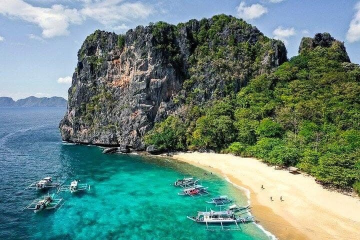 Aerial view of a white-sand beach below a limestone cliff, with bangka boats anchored in turquoise water, on the Tour B route in El Nido
