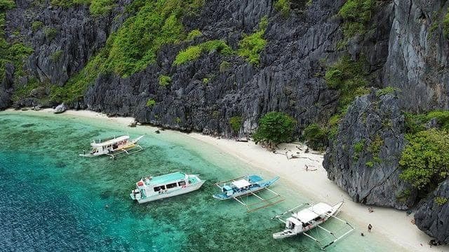 Bangka boats anchored off a narrow white beach at the foot of grey limestone cliffs, on the Tour C route in El Nido