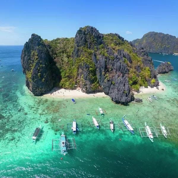 Aerial view of a small limestone island with white beaches on both sides and bangka boats moored over the reef, on the Tour A route in El Nido