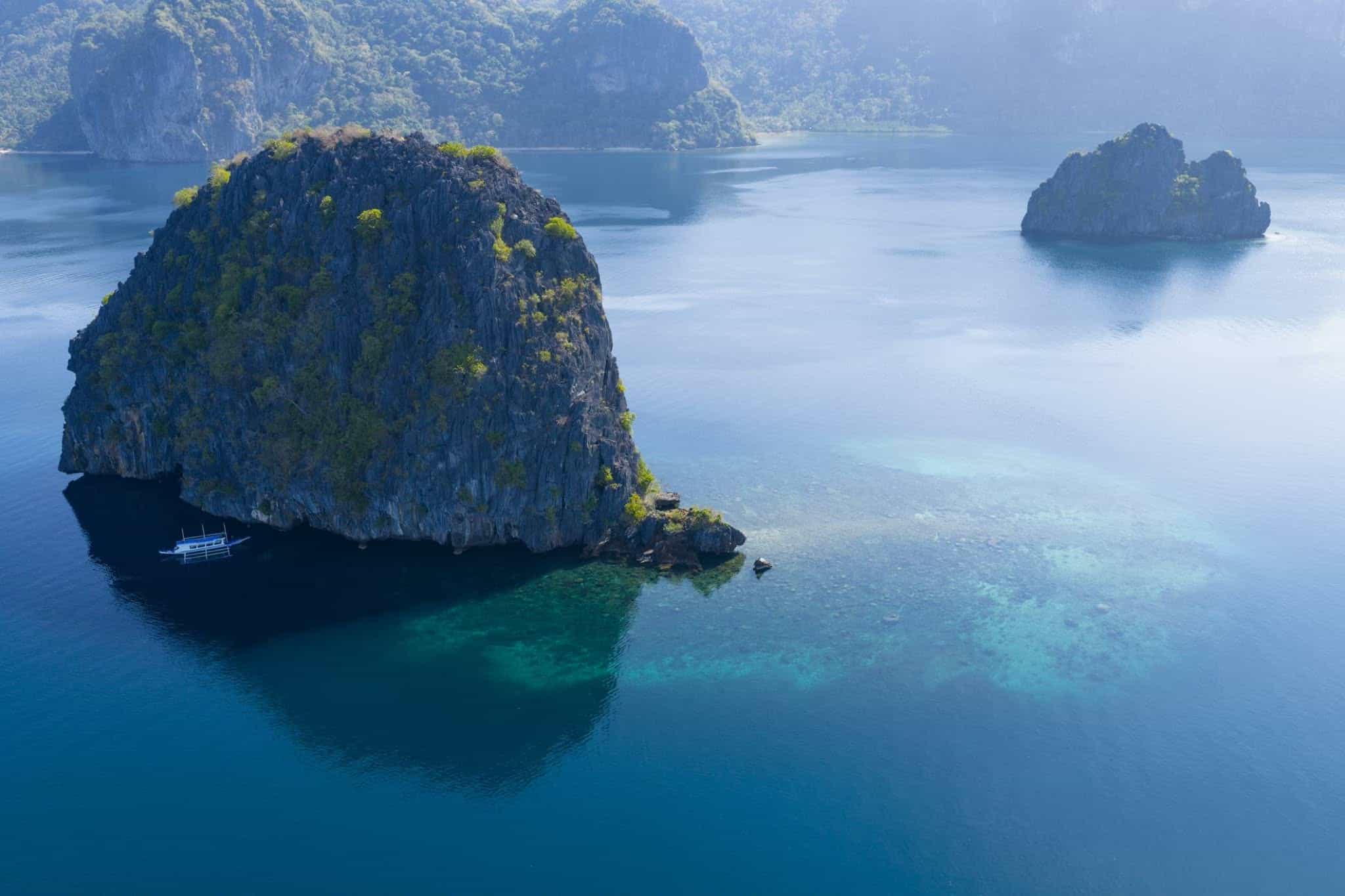 Aerial view of dark limestone islands rising from a calm blue bay, with a single bangka moored in the shadow of a cliff, El Nido