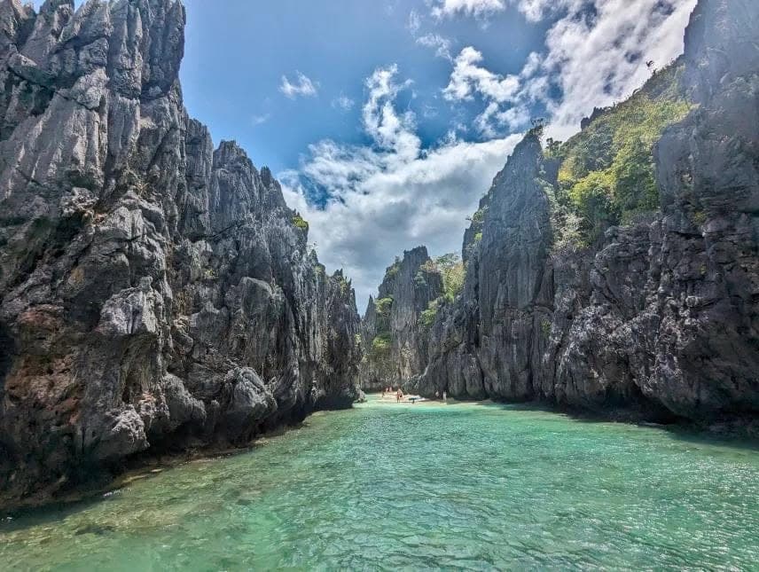Clear green water running between tall grey limestone walls toward a small sandy beach, on the lagoon route in El Nido