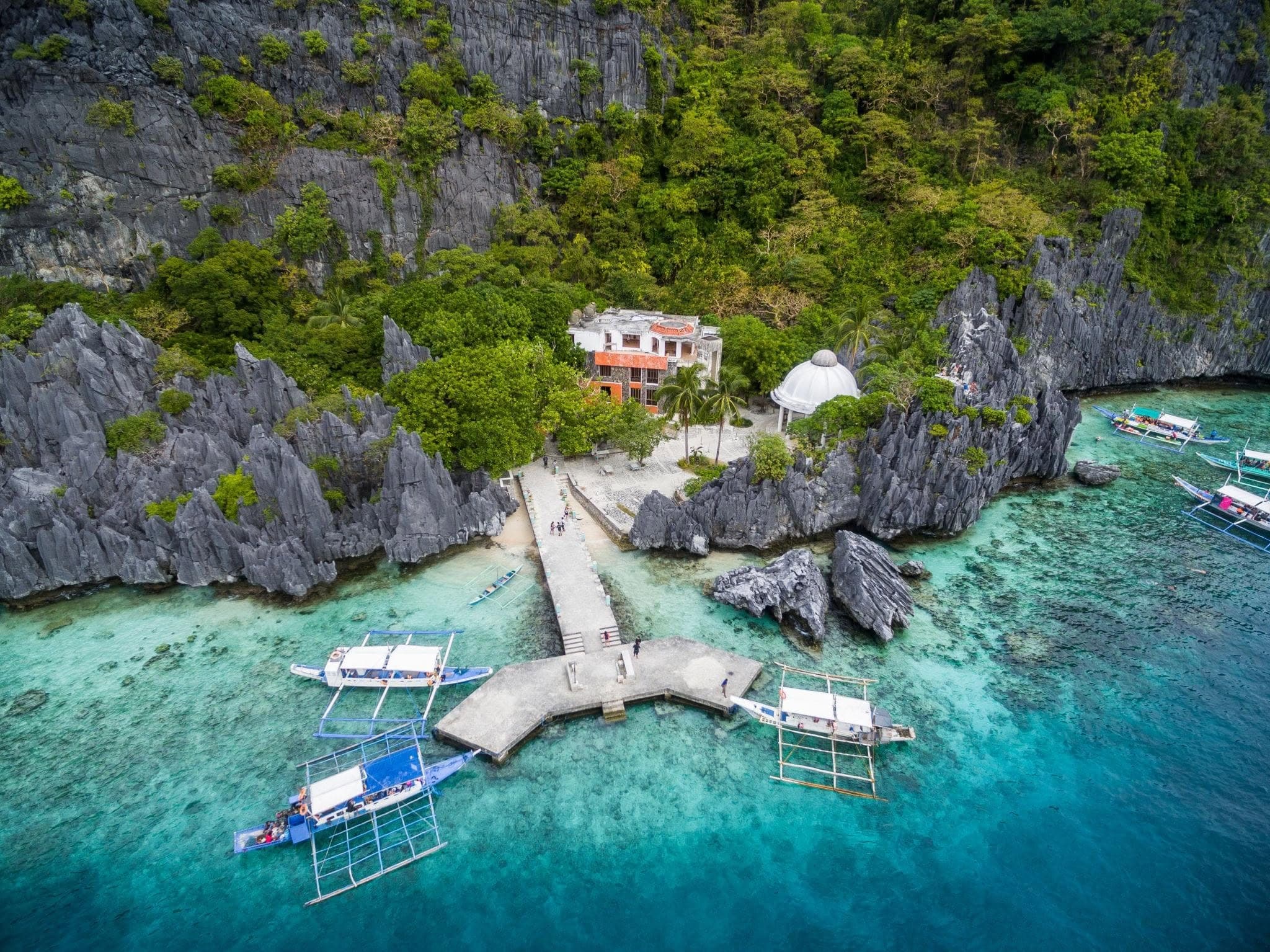 Aerial view of the Matinloc Shrine, a white dome and old building among jagged limestone, with a concrete pier and bangka boats in clear water, El Nido