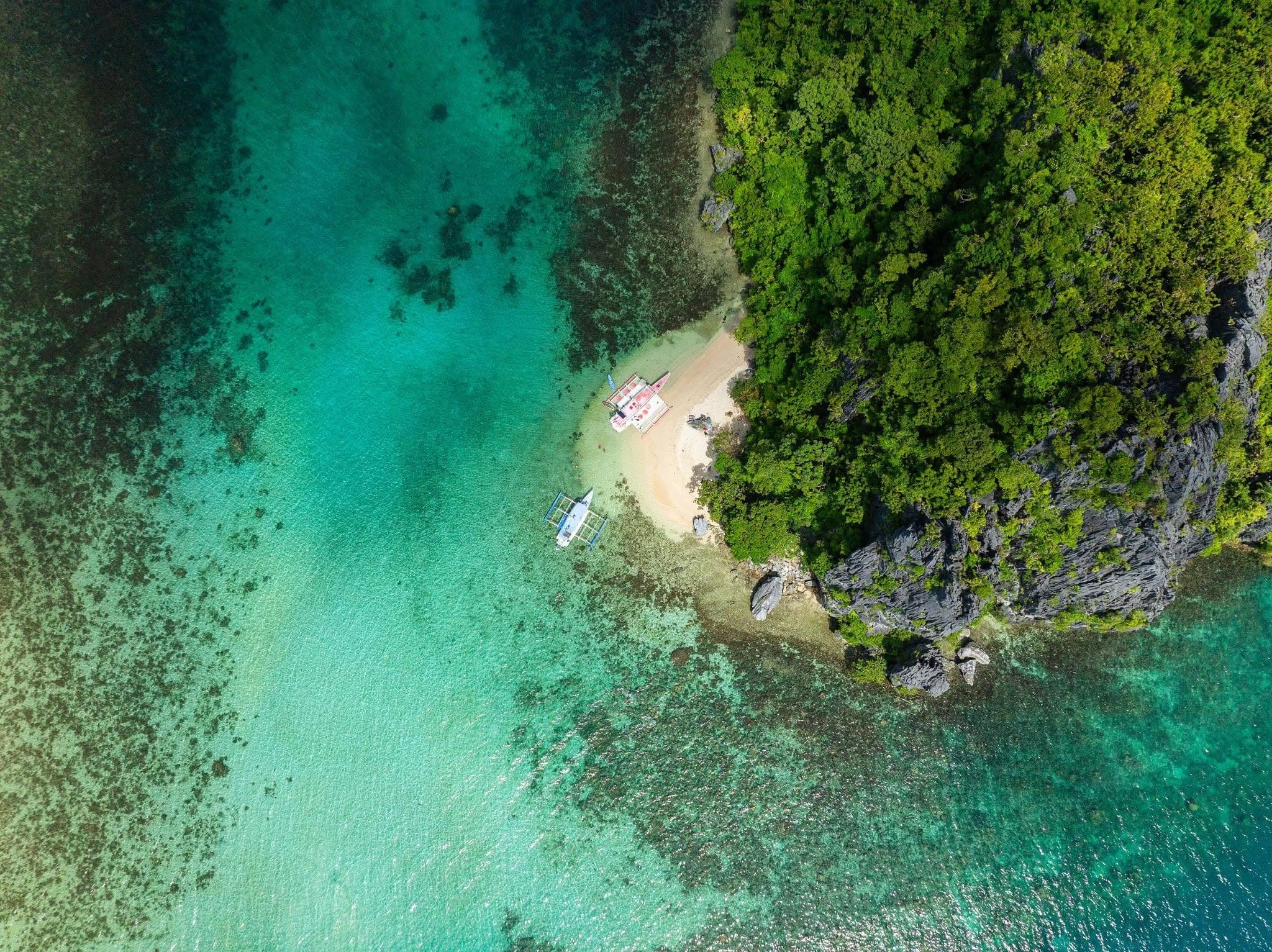 Top-down aerial view of a small sandy beach beside forested limestone, with two bangka boats in clear green reef water, El Nido