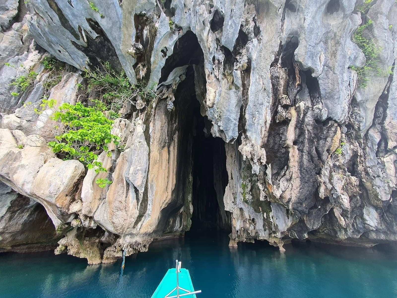Bow of a turquoise bangka nosing toward a tall cave entrance in a limestone cliff, on the Tour B route in El Nido