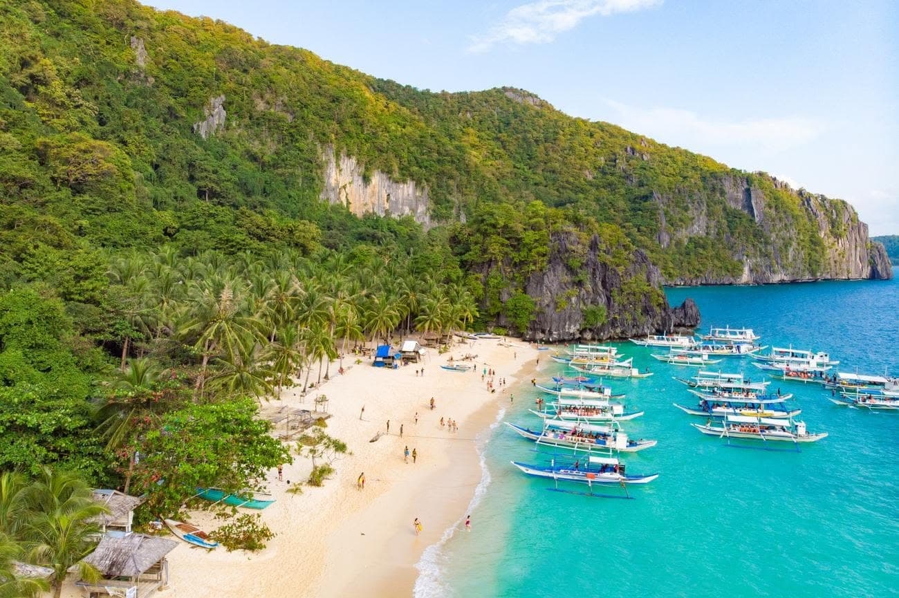 Aerial view of Seven Commando Beach, a long palm-lined white beach with bangka boats anchored in turquoise water below forested cliffs, El Nido